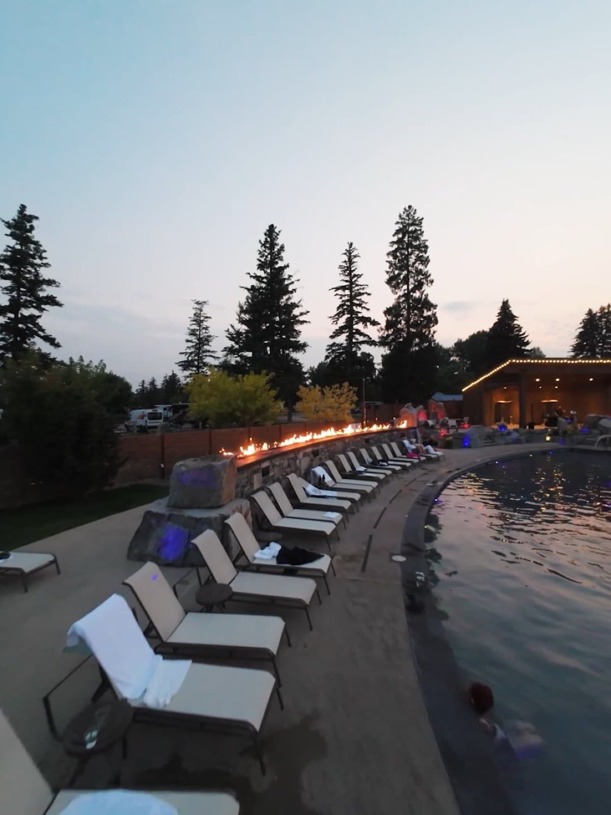 White loungers lined along the lit outdoor pool at Bozeman Hot Springs at dusk, pines behind