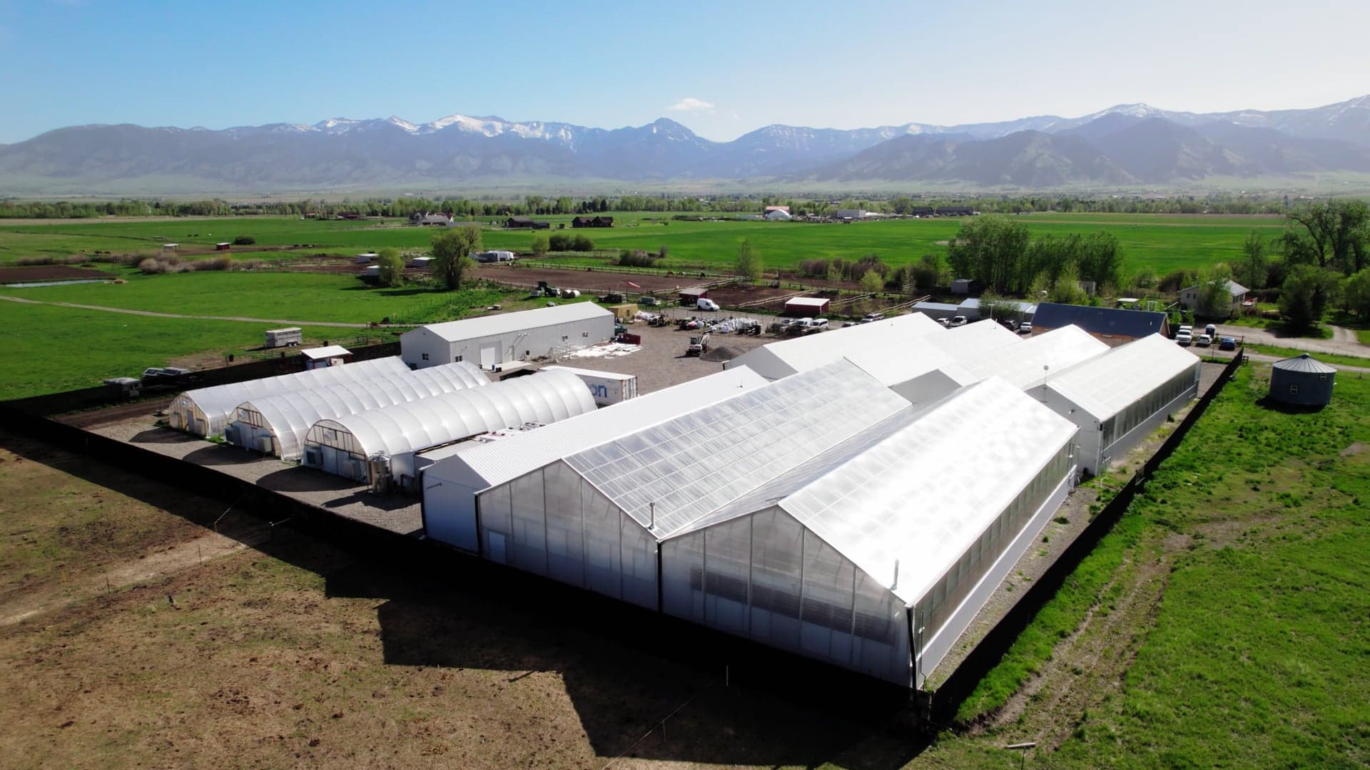 Aerial over Collective Elevation's white greenhouses on a green field with the Bridger range behind