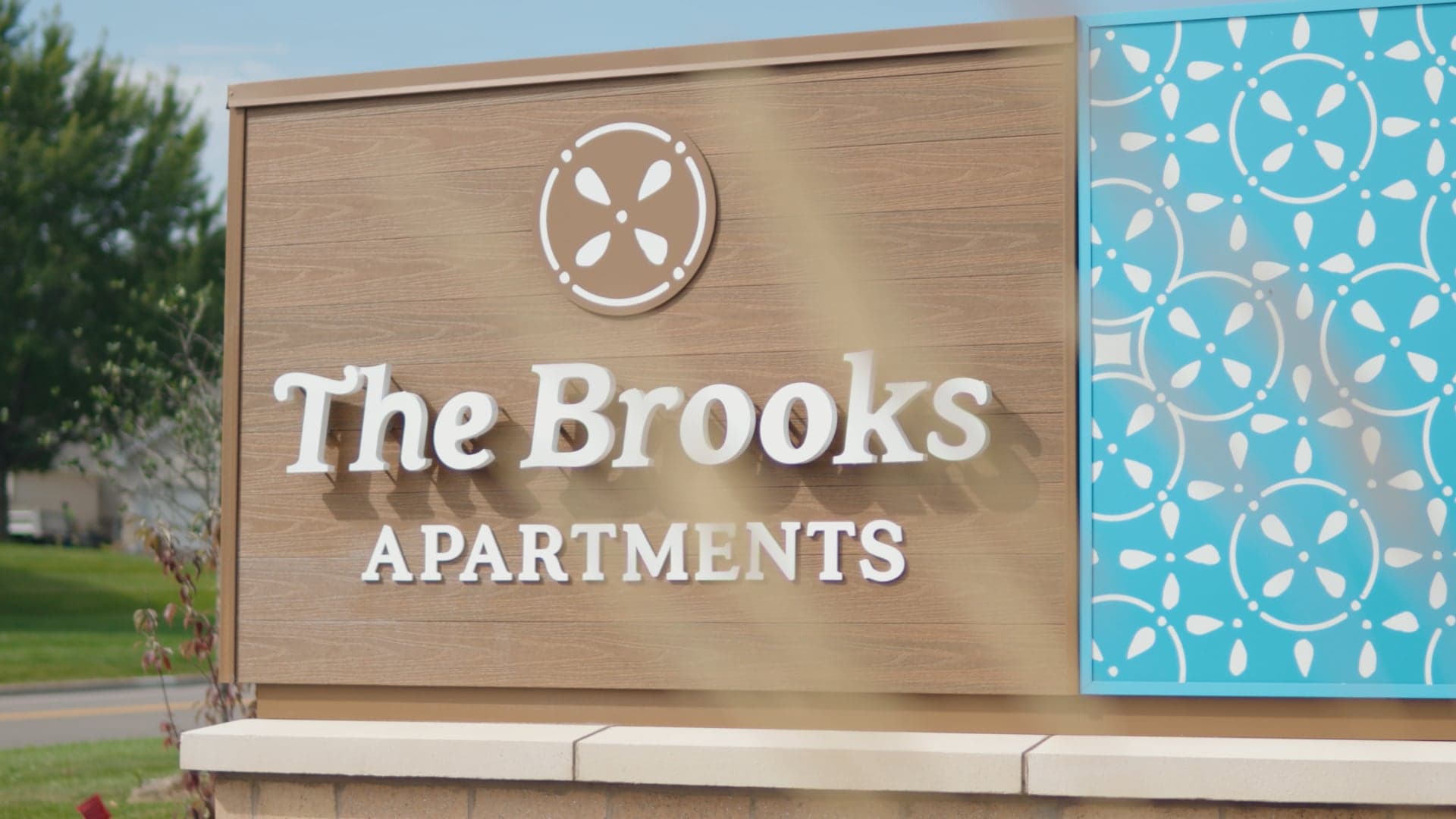 Carved monument sign reading The Brooks Apartments beside a patterned blue panel at the property entrance