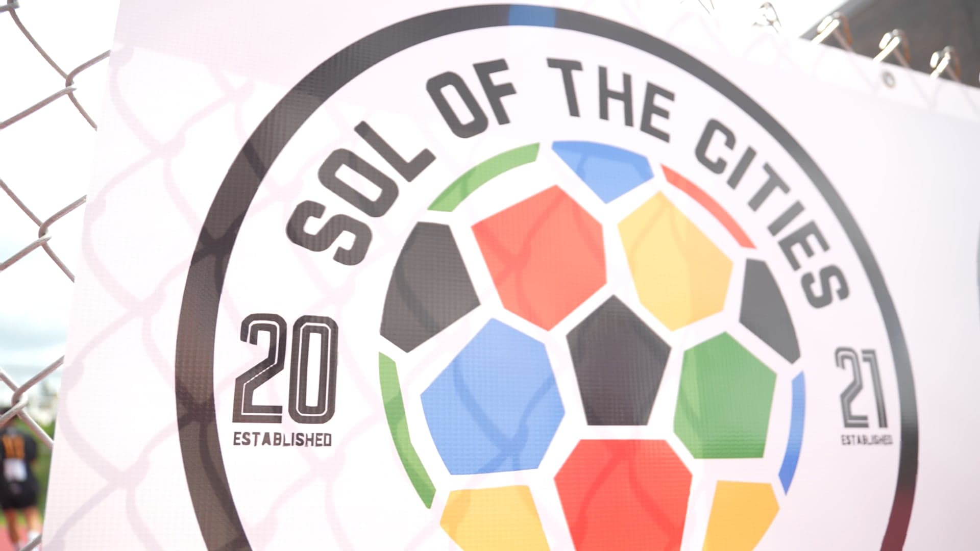 Sol of the Cities crest with a multicoloured soccer ball, printed on a banner against a fence
