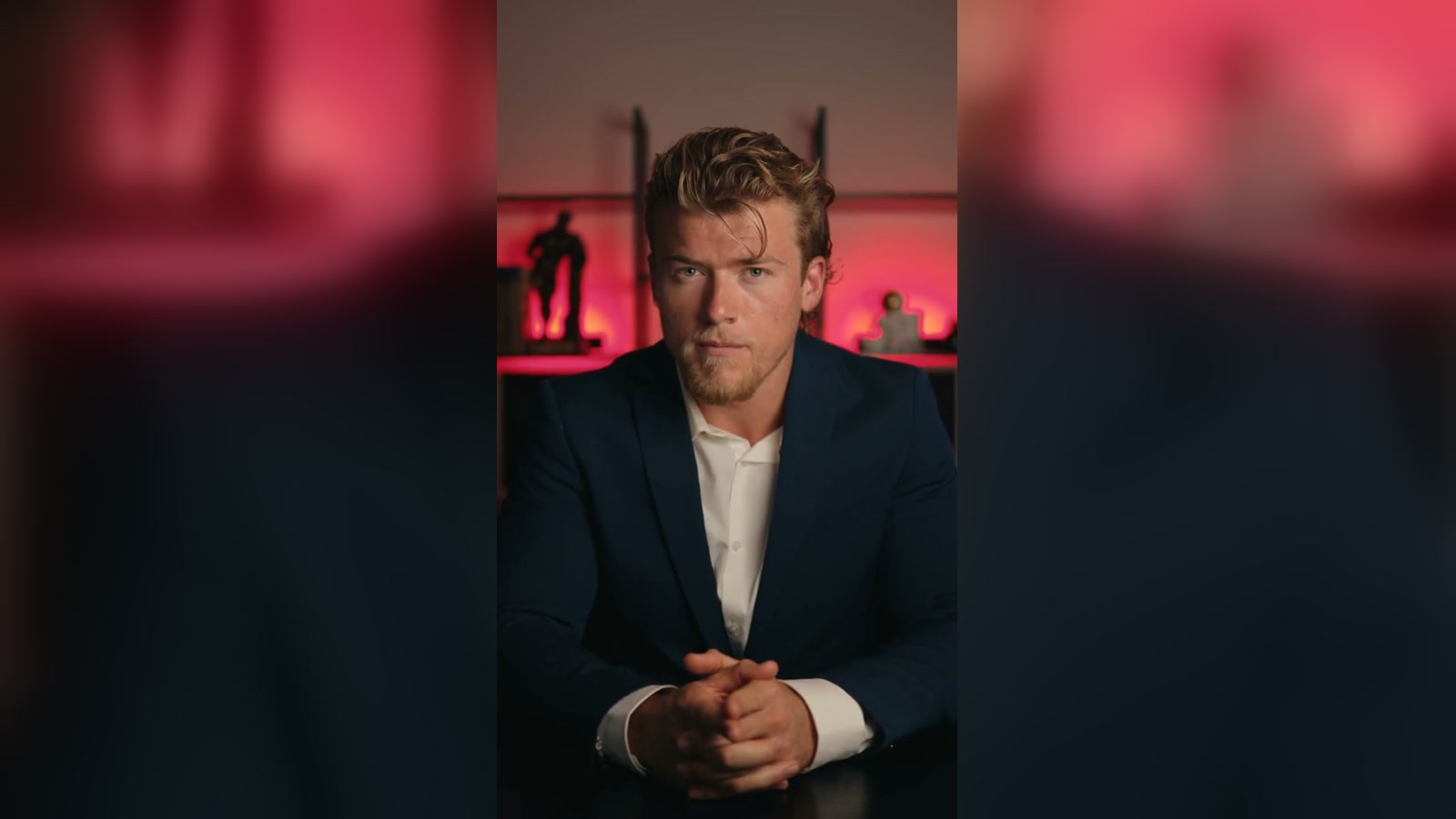 Brandon Clark in a navy blazer, hands clasped, seated in front of a red-lit shelf unit.